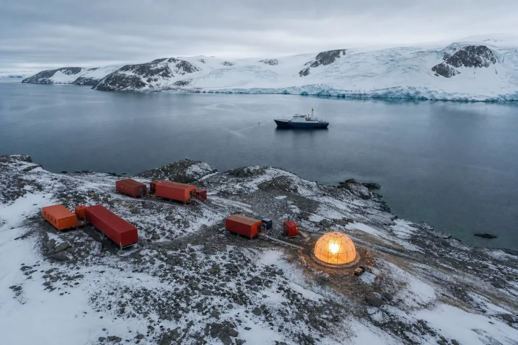 Aerial view of an Antarctic research station on a rocky coast with a clear dome and expedition ship in the bay