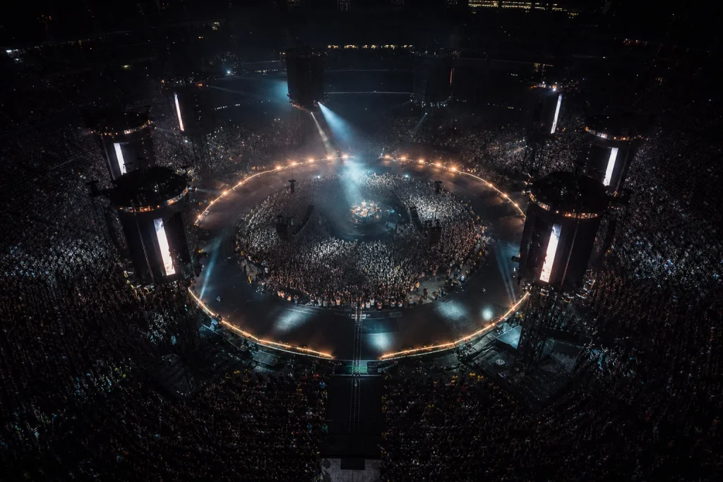 Overhead view of a ring-shaped concert stage with fans inside the center pit and surrounding towers